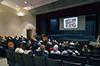 Audience members seated in an audience listen as a speaker describes an image on the screen 